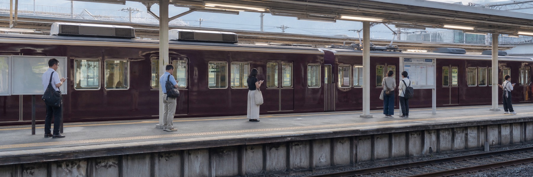 朝の駅ホームで深いえんじ色の電車とスマートフォンを確認する通勤客